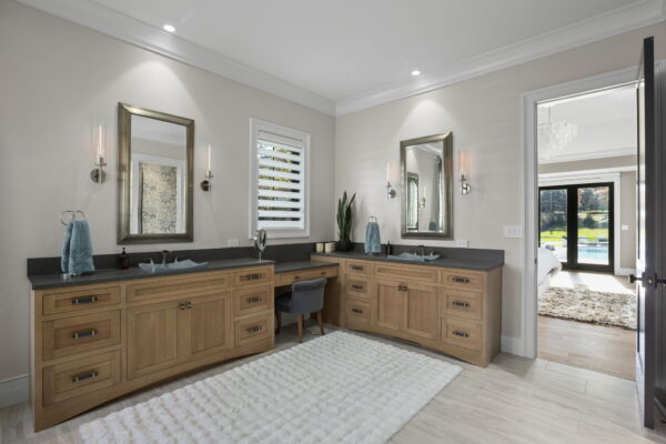 A bathroom with natural wood cabinets, dark stone countertops, light gray tile flooring, and plush rugs.