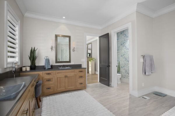 A bathroom with natural wood cabinets, dark stone countertops, light gray tile flooring, and plush rugs.