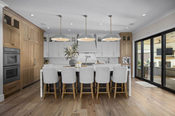 A kitchen with floor-to-ceiling cabinets in white and natural wood, white marble countertops and backsplash, a large island with seating, and a view of the backyard.