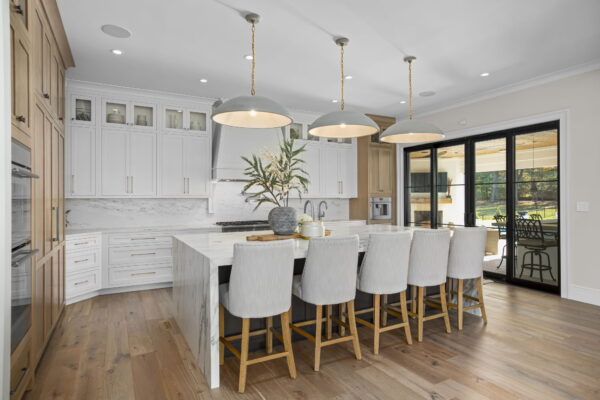 A kitchen with floor-to-ceiling cabinets in white and natural wood, white marble countertops and backsplash, a large island with seating, and a view of the backyard.