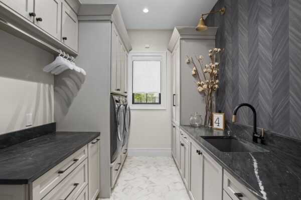 A galley-style laundry space with neutral cabinets, black stone countertops, and neutral floor tile in a herringbone layout.