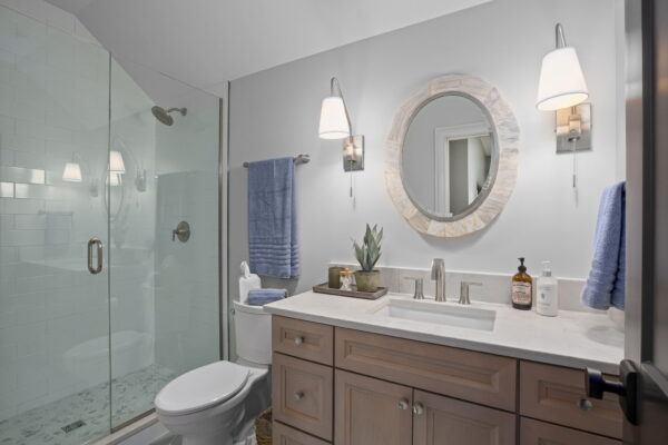 A tiled shower with glass doors next to a single vanity with medium wood cabinets, a light stone countertop, steel fixtures, and bell sconces flanking a circular mirror.