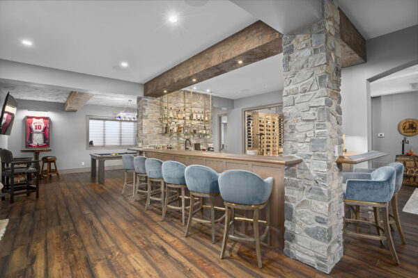 Bar height stools upholstered in light blue suede line the front of a bar with wood and stone finishes.