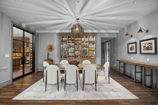 Upholstered dining chairs surround a rectangular dining table in the center of a room with gray walls, rustic wood flooring, and a decorative chandelier.