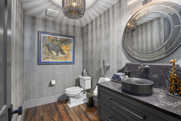 A bathroom with light gray textured wall covering, a standard toilet, a urinal, a single vanity topped with a dark stone countertop and matching vessel sink, and African safari-themed decor.