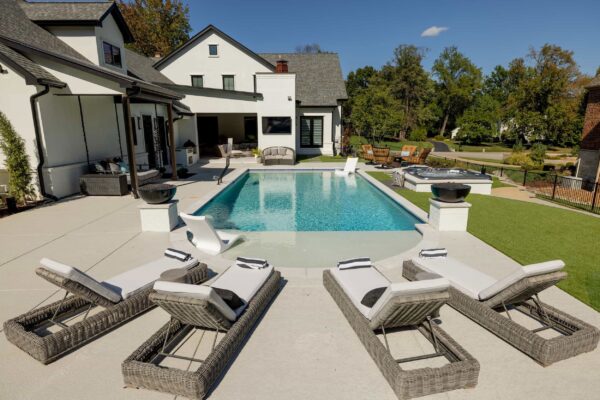 Four wicker lounge chairs with upholstered cushions positioned at the edge of an in-ground beach-entry pool in a residential back yard.