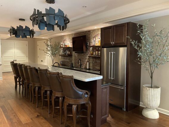 A bar area with dark wood and brick finishes, a stainless steel French door refrigerator, a wall-mounted TV, a polished white stone countertop, and a row of eight chairs upholstered in dark leather.