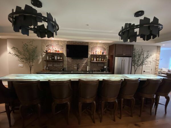 A bar area with dark wood and brick finishes, a stainless steel French door refrigerator, a wall-mounted TV, a polished white stone countertop, and a row of chairs upholstered in dark leather.