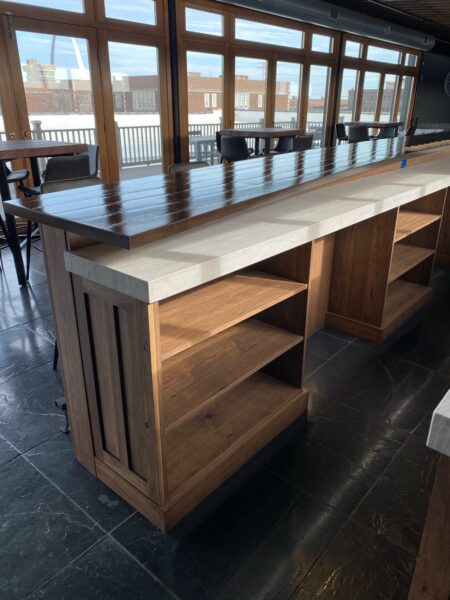 A bar that has a wood base with built-in shelving and a contrasting tiered countertop.