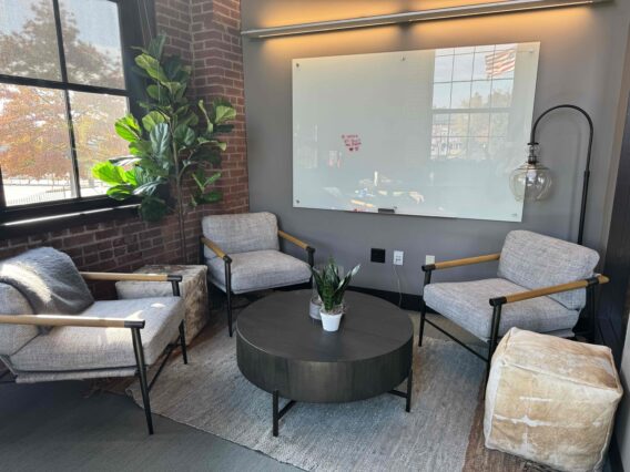 Three gray upholstered armchairs positioned around a low circular table in front of one wall with a whiteboard and another wall with exposed brick and a window.