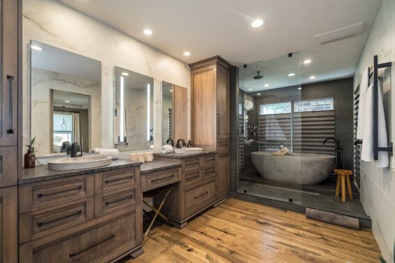 A bathroom with a double vanity and flanked by floor-to-ceiling storage, dark stone countertops, rustic wood casework and flooring, and a walk-in shower and soaking tub combo behind a frameless glass partition.