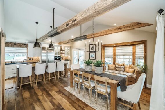 An open-concept kitchen and dining area with rustic wood finishes, exposed brick, and a rectangular dining set placed parallel to a cushioned bay window and perpendicular to an island with six upholstered chairs.