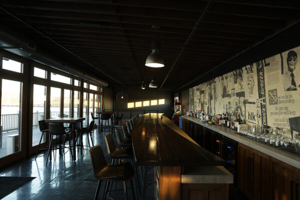 Natural light reflects off a dark bar top that runs the length of a room with black tile flooring, a wall of exterior French doors, and an exposed ceiling.