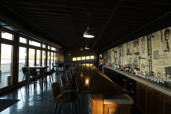 Natural light reflects off a dark stone bar top that runs the length of a room with black tile flooring, a wall of exterior French doors, and an exposed ceiling.