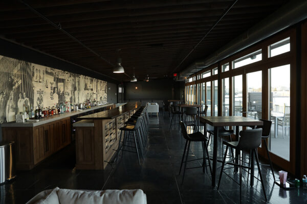 A bar with dark leather chairs runs the length of a room that has black tile flooring, a wall of exterior French doors, and an exposed ceiling.