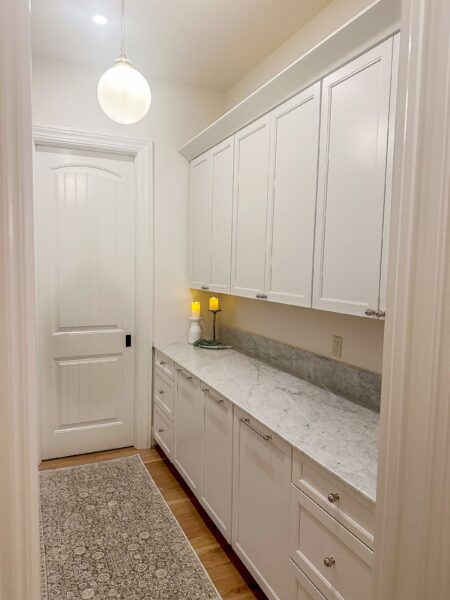 A room with a globe pendant, a linear section of upper and lower cabinets, a white stone countertop, and a neutral floral runner on an oak floor.