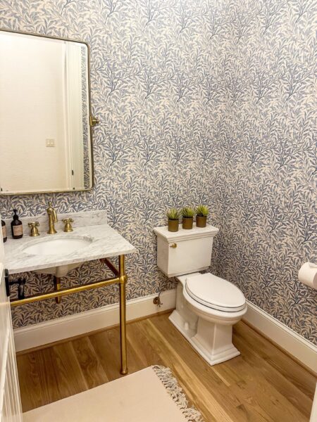 A white stone-topped console sink with brass hardware next to a standard height toilet in a room with oak flooring and a floral motif wallpaper.