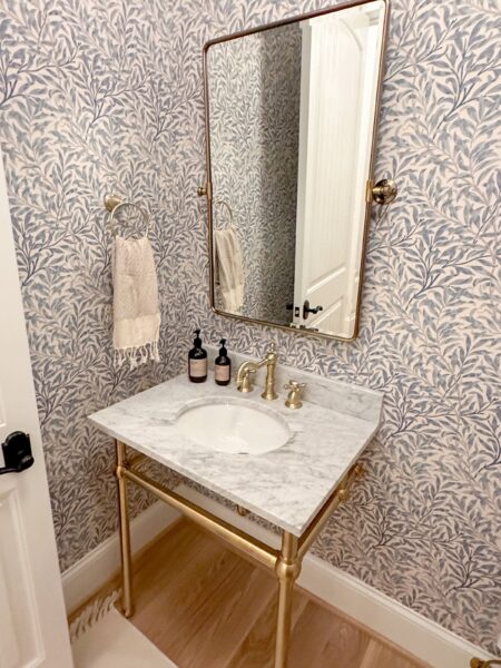 A rectangular mirror hangs above a white stone-topped console sink attached to a wall covered in a floral motif wallpaper.
