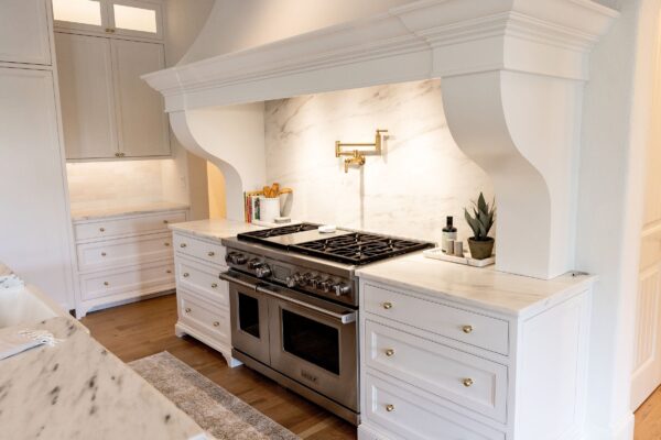 A stainless steel gas range with white marbled stone countertops on each side, a matching backsplash, a brass pot filler faucet, and a white hood with a custom millwork surround.