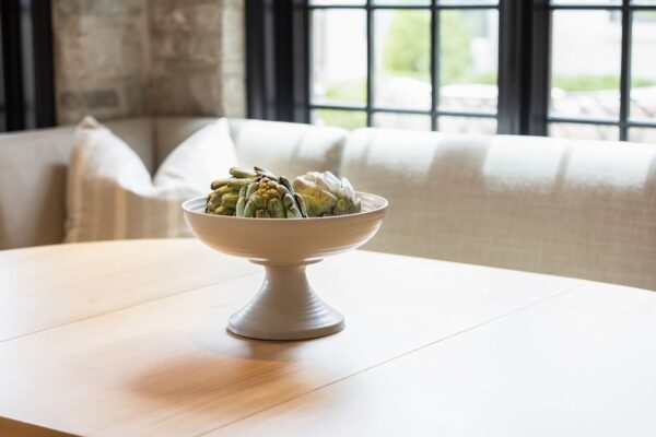 Artichokes inside a white pedestal bowl in the center of an oak table.