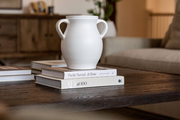 A white ceramic vessel sits on top of a stack of books titled "Pacific Natural At Home" by Jenni Kayne and "AD at 100" by Abrams.
