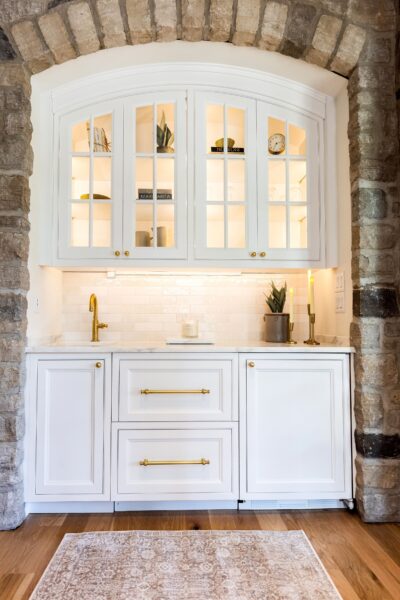 A wet bar built into an arched stone alcove features white cabinets, a white Zellige tile backsplash, a white marble countertop, and brass hardware.