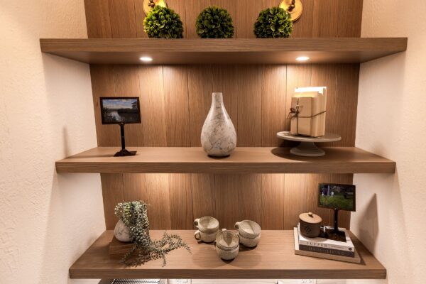 Oak shelves with integrated recessed lighting built into an alcove and styled with artificial plants, stoneware, books, and framed photos.