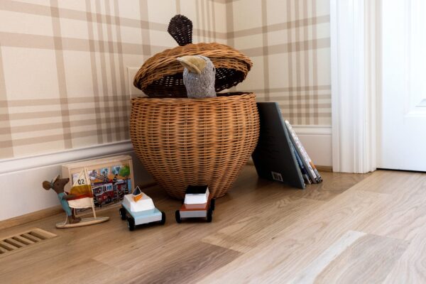 A woven basket that houses children's toys is placed on a white oak floor in front of a neutral plaid wallpaper.
