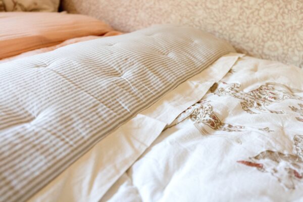 Layers of solid coral, neutral pinstripe, and embroidered bedding against a backdrop of neutral floral motif wallpaper.