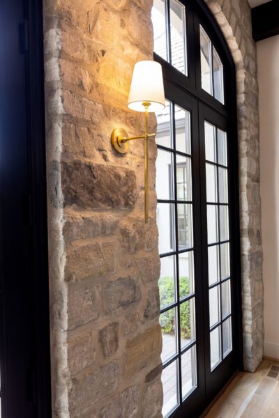 A long-stem brass sconce with a classic white shade installed on a gray stone wall next to a black French double door with a grid of muntins.