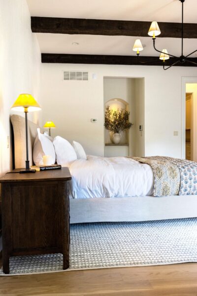 A white and floral motif bed in a gray upholstered bed frame flanked by dark wood nightstands and table lamps and grounded with a low-pile rug.