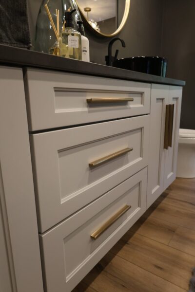 White shaker vanity with dark stone countertop and brass pulls, a black vessel sink under a brass-framed round mirror, and wood‑look plank floor tile.