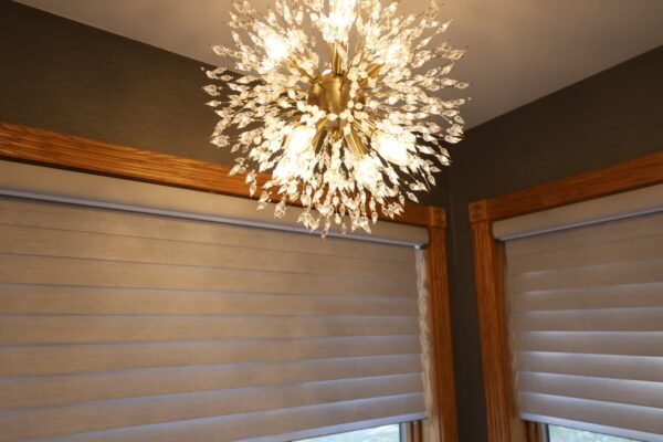 Gold-finish Sputnik-style chandelier with clear crystal droplets hangs above oak-trimmed windows with beige roller shades.