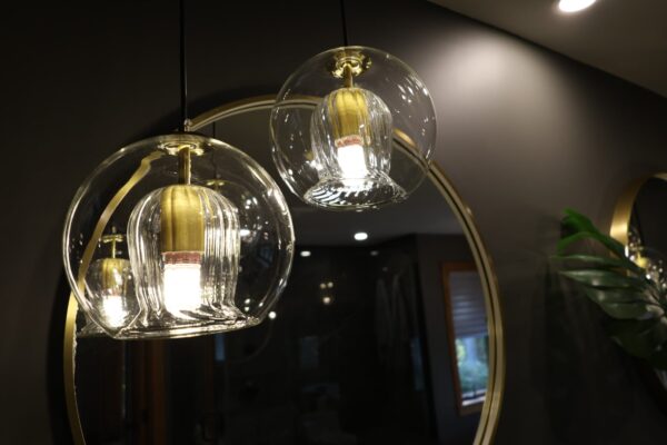 Two ribbed clear‑glass globe pendants with brass fittings cast warm light against a dark grey wall, reflected in a large round brass‑framed mirror.