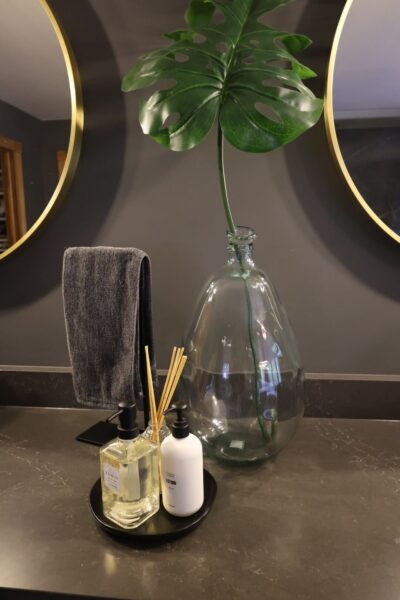 A glass vase with monstera leaf and a tray of bath products on a charcoal quartz vanity countertop beneath gold‑framed mirrors.