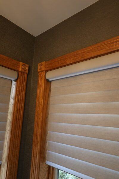 Corner view of two windows framed in honey oak trim with Craftsman rosette blocks, dark grasscloth-textured walls and beige solar roller shades.