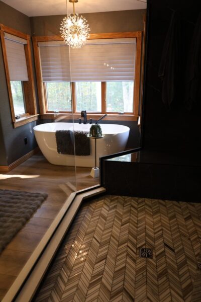 Freestanding white tub beneath oak-framed windows with beige shades, charcoal walls, walk-in glass shower with chevron marble floor tile and matte black fixtures.