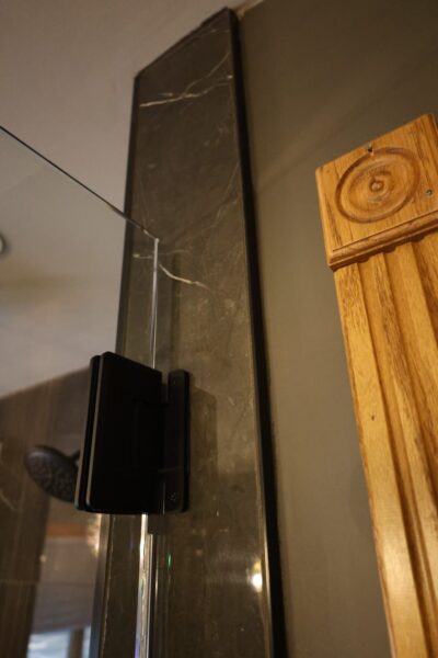 Detail view of glass shower door on matte black hinge attached to black marble wall tile.