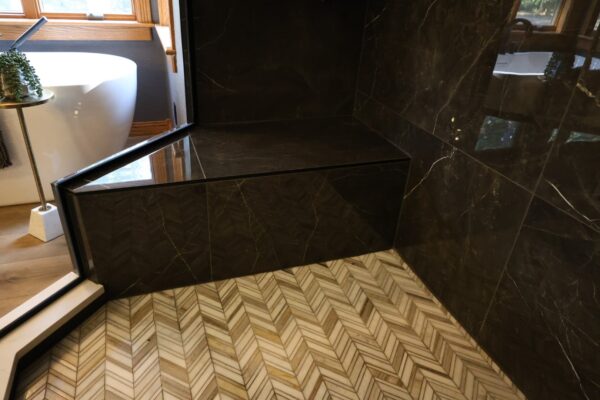 Detail view of a black marble-tiled shower bench and light herringbone patterned floor tiles.