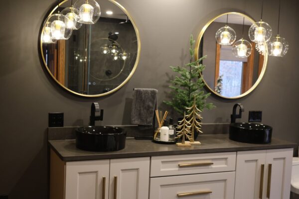Modern white double vanity with brass pulls and charcoal-gray countertop with black vessel sinks, topped by gold round mirrors and glass pendants.