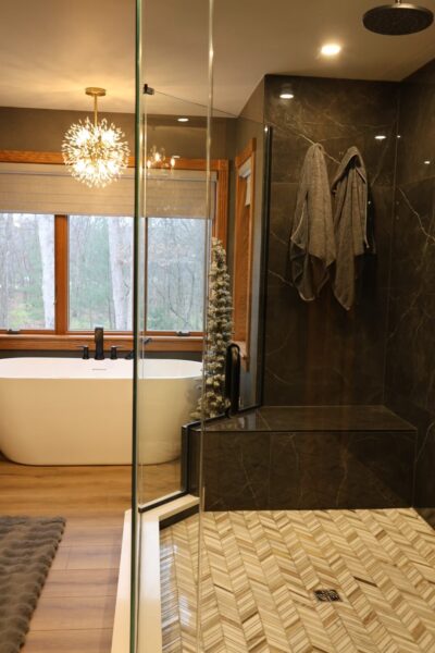 Frameless glass shower with dark marble walls and herringbone marble floor meets freestanding tub with matte black fixtures under oak‑framed window.