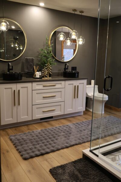 Matte charcoal walls, white shaker vanity with brass pulls, black stone top, round brass mirrors, and a wide-plank wood-look floor.
