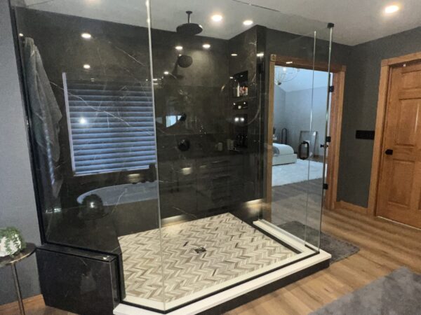 Frameless glass shower clad in black marble with matte-black rain shower, marble bench, and chevron-patterned floor tile.