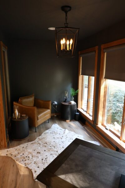 Charcoal walls in an industrial-modern room with oak-framed windows and grey shades, and a cognac leather armchair flanked by two round wood-topped side tables.