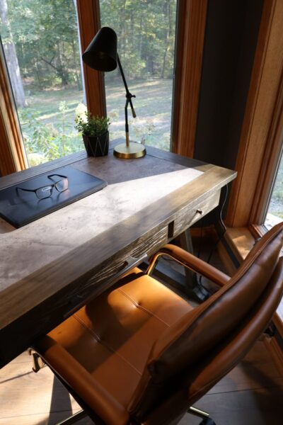 Sunlit home office with grey concrete-look desktop in a dark wood frame, cognac leather swivel chair, brass-accent adjustable lamp, and oak-framed windows.