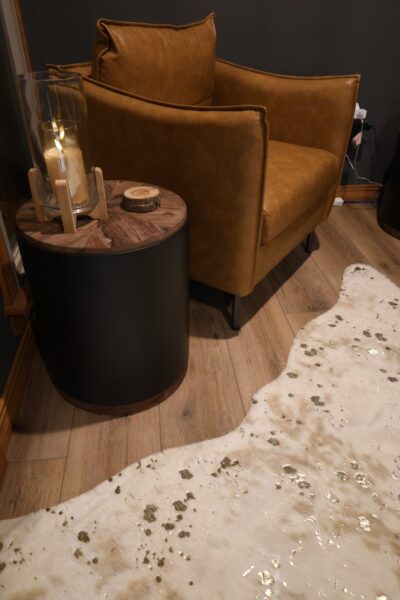 Cognac leather armchair on black metal legs next to a matte-black, wood‑topped side table, on a gold‑flecked white cowhide rug over oak flooring.