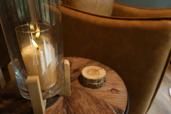 Round wood side table holding a glass cylinder with ivory pillar candle on pine stand and bark-edged coasters next to a caramel leather armchair.