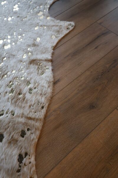 A white faux cowhide rug with gold foil specks on a medium‑brown wide‑plank oak floor.