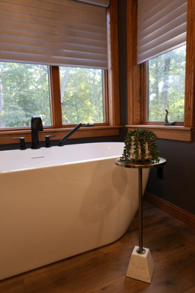 Freestanding white tub with matte black faucet, and a marble‑and‑brass side table with cascading succulent beneath oak-framed windows.